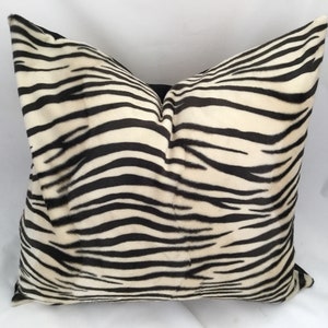 May include: A square decorative pillow with a zebra stripe pattern. The pillow features black stripes on a cream-colored background. The pillow is made of a soft, plush material and is designed for home decor.