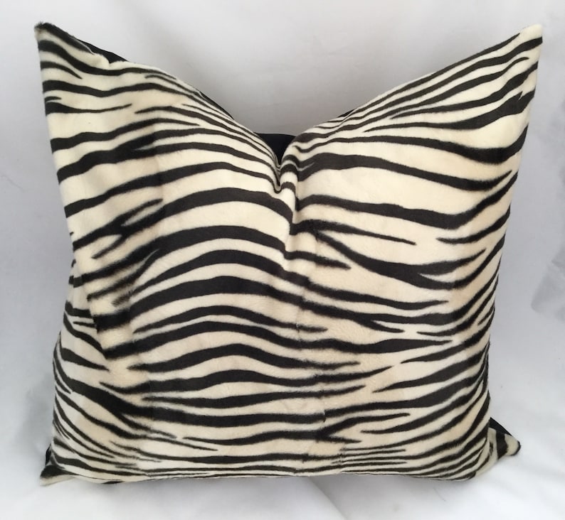 May include: A square decorative pillow with a zebra stripe pattern. The pillow features black stripes on a cream-colored background. The pillow is made of a soft, plush material and is designed for home decor.