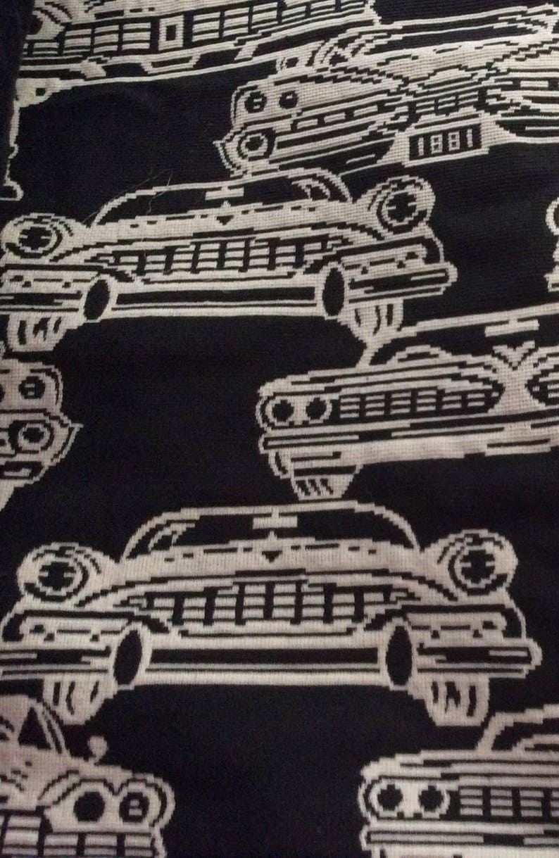 May include: Black and white fabric with a repeating pattern of vintage cars. The cars are stylized with a pixelated design, and some include the number "1881". The overall design is a retro aesthetic.