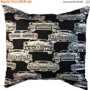 May include: Black decorative pillow with a repeating pattern of white vintage cars. The cars are detailed with headlights, grilles, and other features. The pillow is square-shaped and appears to be made of a soft fabric. The text "1881" is visible on some of the cars.