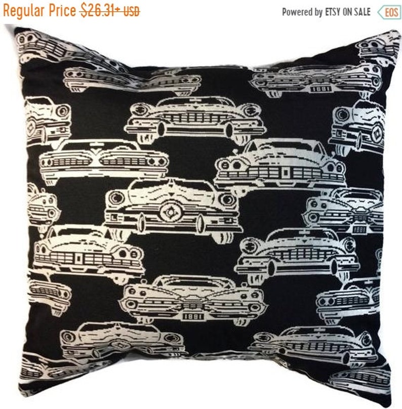 May include: Black decorative pillow with a repeating pattern of white vintage cars. The cars are detailed with headlights, grilles, and other features. The pillow is square-shaped and appears to be made of a soft fabric. The text "1881" is visible on some of the cars.