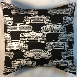 May include: Black throw pillow with a repeating pattern of white vintage cars. The cars are detailed with headlights, grilles, and other features. The pillow is square and appears to be made of a woven fabric.