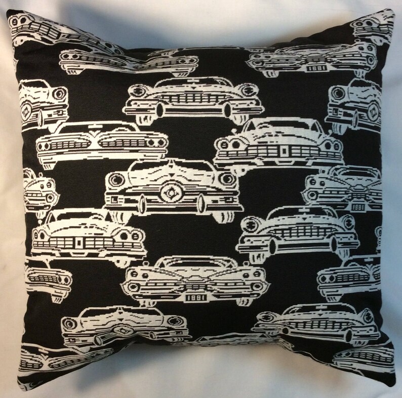 May include: Black throw pillow with a repeating pattern of white vintage cars. The cars are detailed with headlights, grilles, and other features. The pillow is square and appears to be made of a woven fabric.