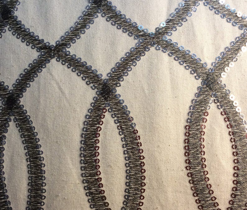May include: Close-up of a white fabric with a silver and brown geometric pattern. The pattern is made of small sequins sewn in a repeating design of circles and diamonds.