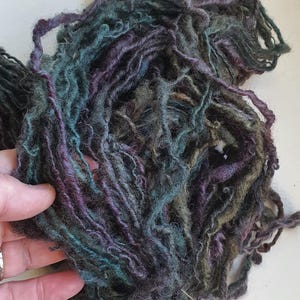 Puede incluir: Un montón de hilo teñido a mano en tonos de verde azulado, morado y verde oliva. El hilo tiene un aspecto texturizado, ligeramente borroso, con hebras de diferente grosor. Los colores se mezclan, creando un aspecto artístico único. Ideal para proyectos de arte textil.