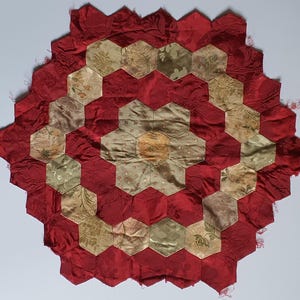 May include: A hexagonal patchwork quilt block featuring a central gold hexagon surrounded by alternating rings of light gold and deep red fabric hexagons. The fabric pieces display various floral and damask patterns with a satin-like finish.