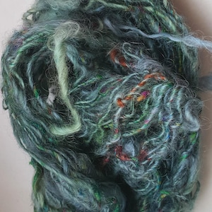 Puede incluir: Ovillo de hilo teñido a mano en tonos turquesa, verde y azul. El hilo tiene un aspecto texturizado, ligeramente difuso, con hebras de color naranja, rojo y motas multicolores. Ideal para proyectos de tejido o ganchillo.