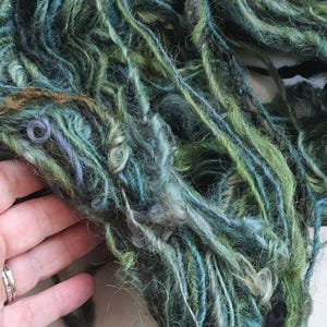 Puede incluir: Primer plano de una madeja de hilo teñido a mano en tonos verdes, azules y un toque de marrón. El hilo tiene un aspecto texturizado y ligeramente difuso, con grosores variables y una mezcla de colores. Ideal para proyectos de tejido o ganchillo.