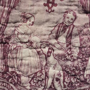 May include: A quilted fabric featuring a maroon and cream toile de Jouy pattern. The design depicts a person in a dress and a person in a vest and trousers sitting outdoors with a dog. The scene includes floral elements and decorative borders.