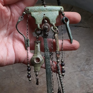 May include: An antique-style pendant necklace with a textured, green-toned metal design. The pendant features various dangling elements, including beads and stylized shapes, suspended from a central, ornate piece. The necklace is held in a hand.