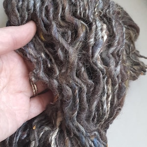 Puede incluir: Primer plano de una madeja de hilo hilado a mano. El hilo es una mezcla de colores marrón oscuro, gris y crema con motas azules y amarillas. El hilo tiene un aspecto grueso y texturizado, ideal para proyectos de tejido o ganchillo.