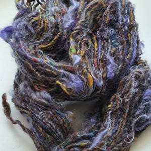 Puede incluir: Primer plano de una madeja de hilo teñido a mano. El hilo es una mezcla de colores, incluyendo tonos de morado, marrón, amarillo y verde. La textura es gruesa y texturizada, con un aspecto ligeramente borroso. Este hilo es adecuado para proyectos de tejido o ganchillo.