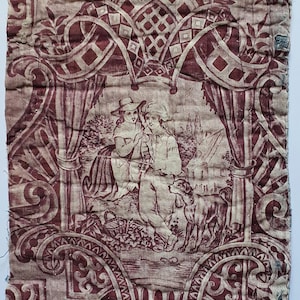 May include: A rectangular piece of quilted fabric featuring a maroon and cream toile de Jouy pattern. The design depicts two figures in period clothing sitting outdoors with a dog, surrounded by ornate floral motifs, drapery, and lattice elements.