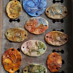 May include: Ten ceramic oval-shaped pendants with a reindeer design. The pendants are painted in various colors, including blue, gold, brown, and green. Each pendant has two holes for hanging.