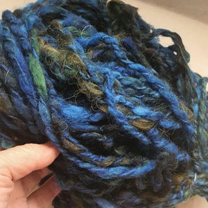 Puede incluir: Primer plano de una madeja de hilo hilado a mano. El hilo es una mezcla de azules, verdes y marrones, creando una apariencia texturizada y orgánica. Las fibras son gruesas e irregulares, lo que sugiere una calidad rústica y artesanal. Ideal para tejer u otros proyectos de arte textil.