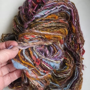 Puede incluir: Ovillo de lana teñida a mano con una mezcla de texturas y colores. La lana presenta tonos marrones, morados, naranjas y azules. La fibra se mantiene unida con un hilo fino de color natural. Ideal para tejer y otras artes textiles.