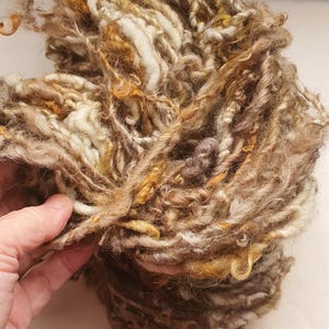 Puede incluir: Primer plano de un ovillo de hilo grueso y texturizado en tonos marrón, beige y dorado. El hilo parece estar hilado a mano, con grosores variables y un aspecto ligeramente rústico. Ideal para tejer u otras manualidades con fibra.