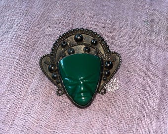 May include: A vintage brooch featuring a green face with detailed features, set within an ornate, aged metal frame. The brooch has a dark, antique finish, and the face is a vibrant, glossy green. The design is reminiscent of ancient art.