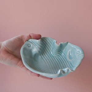 May include: A light blue ceramic fish-shaped bowl with a wavy design. The bowl has a textured surface with a fish face on each end. The bowl is held in a hand against a pink background.