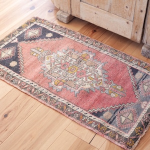 May include: A rectangular area rug featuring a central pink field with a geometric medallion in shades of cream, yellow, and grey. The rug is framed by dark grey borders with repeating diamond motifs and intricate patterns along the edges.