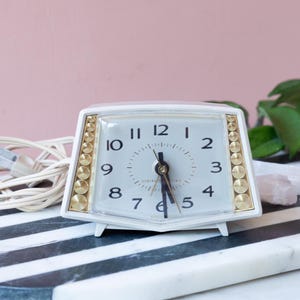 May include: A vintage white electric alarm clock with gold accents. The clock has a rectangular face with black numerals and hands. The clock is on a black and white striped surface.