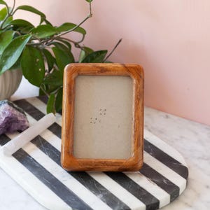 May include: A rectangular wooden picture frame with a light brown finish. The frame is empty and sits on a black and white striped tray. A purple crystal and a white crystal are also on the tray, with a white ceramic pot with green leaves in the background.