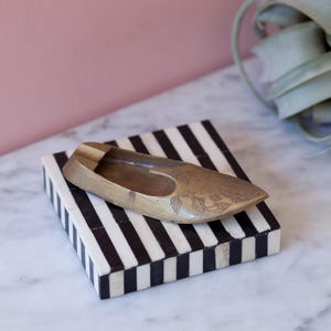 May include: A brass ashtray shaped like a shoe with an intricate design sits on a black and white striped tray.