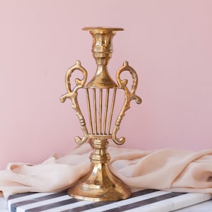 May include: A gold metal candlestick with a lyre design. The candlestick has a round base and a tall, slender body with a decorative top.