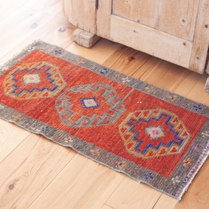 May include: A rectangular woven rug featuring a vibrant orange center field with three repeating geometric medallions in shades of blue, yellow, and pink. The rug is framed by a wide, textured gray border with small colorful accents and fringed edges.