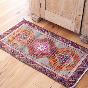 May include: A rectangular area rug featuring a gray background with three central geometric medallions in vibrant orange and hot pink. The rug is framed by a dark purple border with intricate patterns and has short white fringe on the ends.