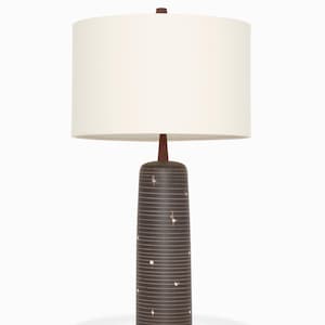 May include: A brown table lamp with a cylindrical base and a white lampshade. The base features horizontal lines and small, scattered white dots. The lamp has a wooden stem and a dark brown finial.