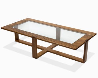 May include: A rectangular coffee table with a clear glass top and a wooden frame. The frame is made of light brown wood and features an X-shaped support in the center. The table is designed with a modern aesthetic.