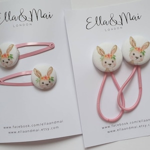 May include: Two pink hair clips and two pink hair ties with white fabric bunny buttons. The buttons have a floral design and a brown bunny face. The text "Ella & Mai London" is printed on the cards.