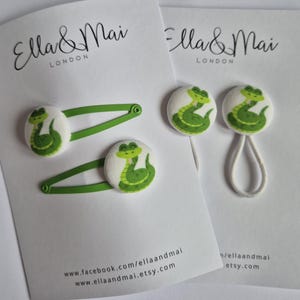 May include: Two sets of hair accessories with a green alligator design on white fabric. One set includes two green hair slides, and the other includes two hair ties. The accessories are on white cards with the text "Ella & Mai LONDON".