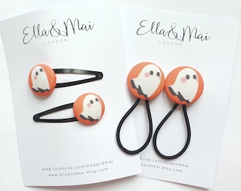 May include: Two sets of hair accessories: barrettes and hair ties. Each accessory features a round orange button with a white ghost illustration. The barrettes have black metal clips, and the hair ties have black elastic bands. The brand name "Ella & Mai London" is visible.