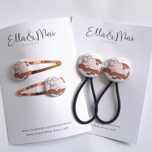 May include: Two sets of pig hair clips and pig hair scrunchies. The hair clips are gold with a pink pig design. The scrunchies are black with a pink pig design. The packaging includes the text "Ella & Mai London".