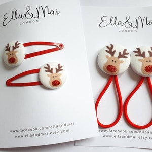 May include: Two sets of festive hair accessories. One set includes red hair slides with reindeer-themed buttons. The other set has red elastic hair ties with matching reindeer buttons. The reindeer have brown antlers and red glitter noses.