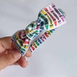 May include: A white headband with a bow featuring a colorful crayon pattern. The crayons are in various colors, including red, yellow, green, and purple. The headband is held by a hand against a white background.