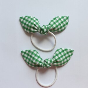 May include: Two green and white gingham hair ties with white elastic bands. The hair ties are tied in a bow shape.
