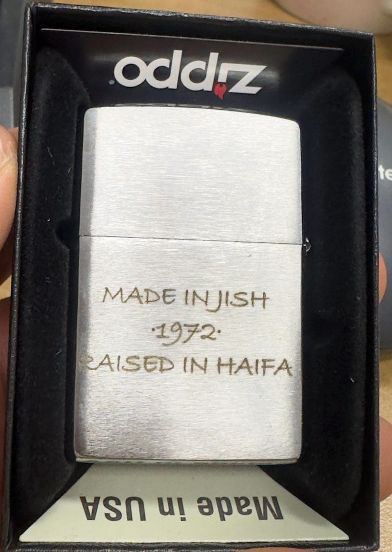 Puede incluir: Un encendedor plateado con el texto "MADE IN JISH 1972 RAISED IN HAIFA" grabado en la parte delantera. El encendedor est&aacute; en una caja negra con el nombre de la marca "oddz" impreso en la tapa. La parte inferior de la caja dice "Made in USA".