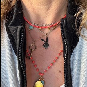 May include: A layered necklace with a variety of charms, including a key, a house, and a bunny. The necklace is made of gold-toned metal and features coral-colored beads.