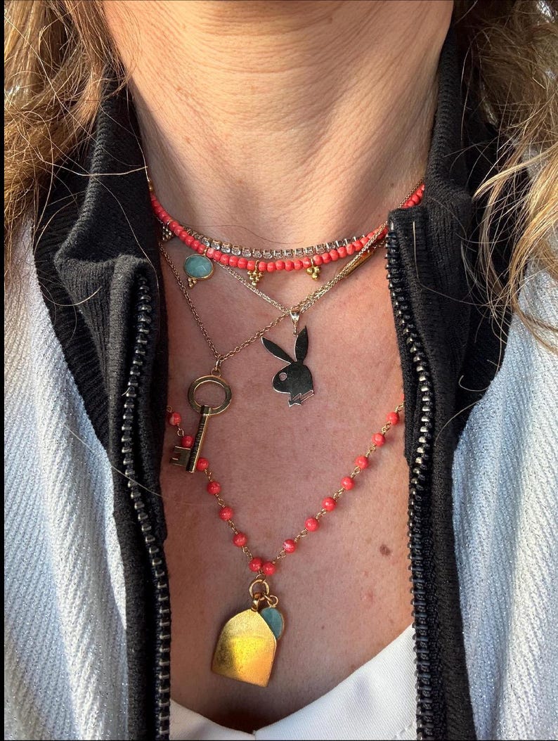 May include: A layered necklace with a variety of charms, including a key, a house, and a bunny. The necklace is made of gold-toned metal and features coral-colored beads.