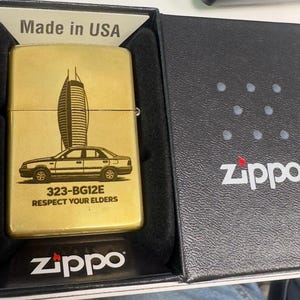 Puede incluir: Un encendedor Zippo dorado con un dise&ntilde;o de coche y rascacielos. El encendedor est&aacute; en su caja negra original, con el logotipo de Zippo visible. El texto "Made in USA" tambi&eacute;n es visible.