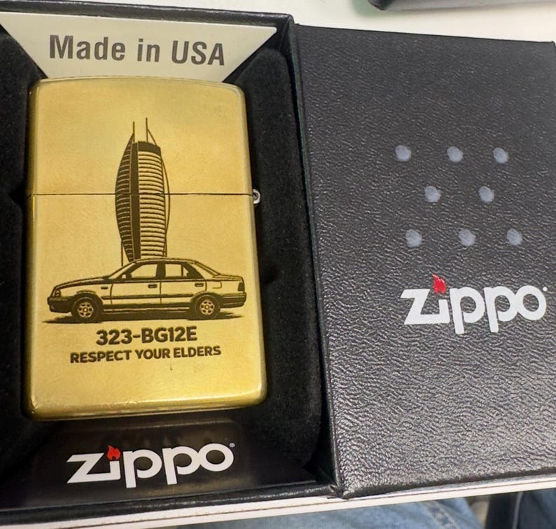 Puede incluir: Un encendedor Zippo dorado con un dise&ntilde;o de coche y rascacielos. El encendedor est&aacute; en su caja negra original, con el logotipo de Zippo visible. El texto "Made in USA" tambi&eacute;n es visible.