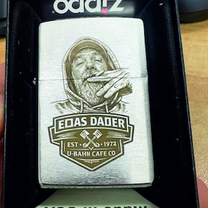 Puede incluir: Un encendedor Zippo plateado con un grabado detallado de una persona fumando, con una sudadera con capucha. El encendedor presenta el texto "EOAS DADER EST. 1972 U-BAHN CAFE CO." y se presenta en una caja negra. La caja dice "Made in USA".