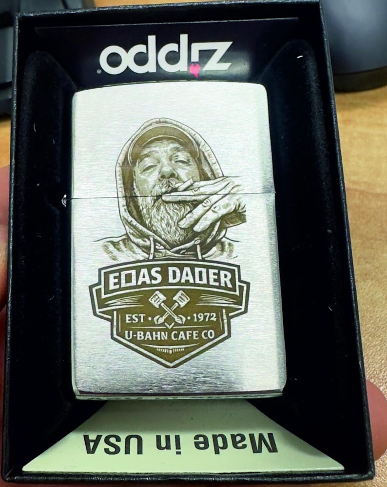 Puede incluir: Un encendedor Zippo plateado con un grabado detallado de una persona fumando, con una sudadera con capucha. El encendedor presenta el texto "EOAS DADER EST. 1972 U-BAHN CAFE CO." y se presenta en una caja negra. La caja dice "Made in USA".
