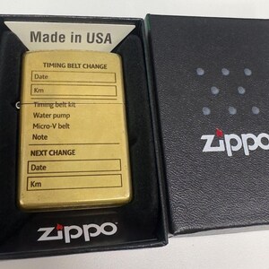 Puede incluir: Un encendedor Zippo dorado en una caja negra. El encendedor tiene una secci&oacute;n para registrar informaci&oacute;n sobre el cambio de la correa de distribuci&oacute;n. La caja es negra con el logotipo de Zippo. El encendedor est&aacute; marcado "Made in USA".