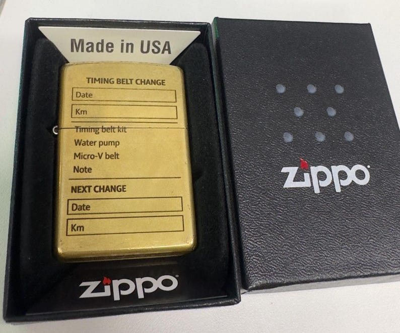 Puede incluir: Un encendedor Zippo dorado en una caja negra. El encendedor tiene una secci&oacute;n para registrar informaci&oacute;n sobre el cambio de la correa de distribuci&oacute;n. La caja es negra con el logotipo de Zippo. El encendedor est&aacute; marcado "Made in USA".