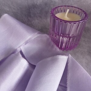 May include: A lavender-colored, ribbed glass votive candle holder with a lit white candle sits next to a folded lavender fabric. The fabric has a soft, silky appearance. The background is a muted gray.