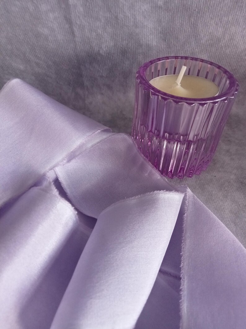May include: A lavender-colored, ribbed glass votive candle holder with a lit white candle sits next to a folded lavender fabric. The fabric has a soft, silky appearance. The background is a muted gray.